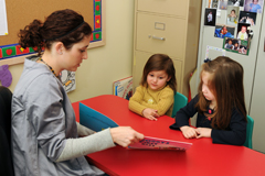 An adult shows a book to two children seated at a red table.